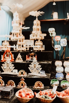 The image depicts a collection of intricate wooden Christmas decorations arranged on black shelves. Among the decorations are large, multi-tiered candle pyramids adorned with small figures and candles. Various other holiday ornaments, such as wooden trees and small nativity scenes, are displayed alongside colorful ceramic candle holders with festive designs. Baskets filled with red and green items are positioned at the bottom of the display.
