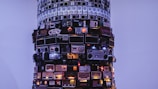 A tall, cylindrical structure is composed of various stacked vintage radios against a pale blue background. The radios, differing in size, color, and design, form a visually striking sculpture with some buttons and dials illuminated, adding a warm glow.