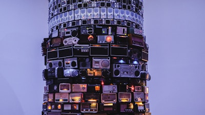A tall, cylindrical structure is composed of various stacked vintage radios against a pale blue background. The radios, differing in size, color, and design, form a visually striking sculpture with some buttons and dials illuminated, adding a warm glow.