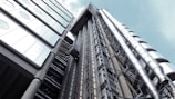 A modern, industrial building facade featuring exposed metallic pipes and large glass windows. The design includes sleek, cylindrical structures aligned vertically, contributing to a futuristic appearance.