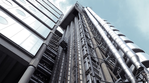 A modern, industrial building facade featuring exposed metallic pipes and large glass windows. The design includes sleek, cylindrical structures aligned vertically, contributing to a futuristic appearance.