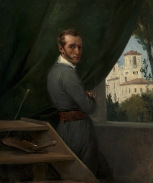 A man in period clothing stands by an open window, holding a paintbrush. He gazes directly at the viewer with a serious expression. Behind him, a lush landscape with a large building or villa is visible. An easel and painter's palette are present in the foreground, suggesting an artist in his studio.