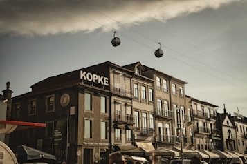 A street scene featuring a row of traditional European buildings with detailed architecture and a prominent sign reading 'Kopke'. Above the buildings, two gondola-like cable cars traverse a wire against a cloudy sky. Small shops and cafes with outdoor seating line the street, adding a lively atmosphere.