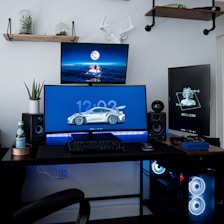 A modern computer setup featuring three monitors on a desk. The main monitor displays a car image with a digital clock. The secondary monitors display digital art and a scenic night image with a full moon. The desk is organized with a keyboard, mouse, gaming controller, and speakers. The wall has decorative elements, including a shelf with small plants and a mounted sculpture of a deer head. LED lighting under the monitors and a gaming PC with illuminated fans add a techy atmosphere.