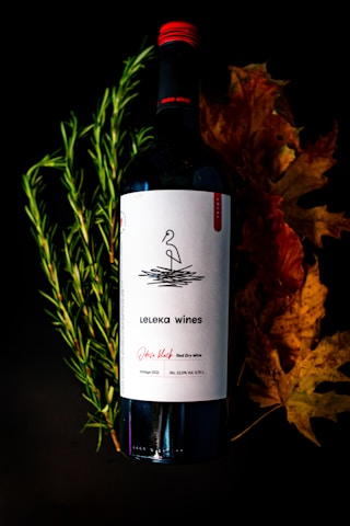 A bottle of red wine labeled 'Leleka wines' is positioned against a dark background. The label includes a simple black and white design with minimalist line art. Surrounding the bottle are fresh green rosemary sprigs on the left and dried autumn leaves on the right, adding a natural and seasonal touch to the composition.