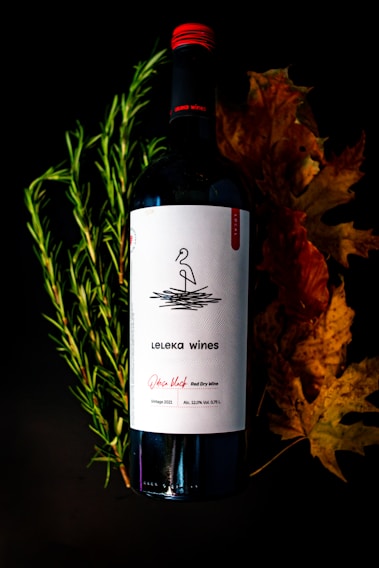 A bottle of red wine labeled 'Leleka wines' is positioned against a dark background. The label includes a simple black and white design with minimalist line art. Surrounding the bottle are fresh green rosemary sprigs on the left and dried autumn leaves on the right, adding a natural and seasonal touch to the composition.
