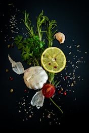 A composition of fresh ingredients, including a whole garlic bulb, a lemon slice, rosemary sprigs, parsley leaves, a small tomato, and scattered peppercorns and salt on a dark background.