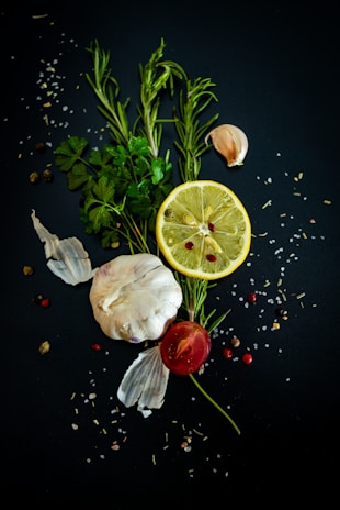 A composition of fresh ingredients, including a whole garlic bulb, a lemon slice, rosemary sprigs, parsley leaves, a small tomato, and scattered peppercorns and salt on a dark background.