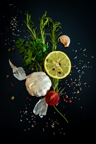 A composition of fresh ingredients, including a whole garlic bulb, a lemon slice, rosemary sprigs, parsley leaves, a small tomato, and scattered peppercorns and salt on a dark background.
