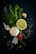 A composition of fresh ingredients, including a whole garlic bulb, a lemon slice, rosemary sprigs, parsley leaves, a small tomato, and scattered peppercorns and salt on a dark background.