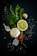 A composition of fresh ingredients, including a whole garlic bulb, a lemon slice, rosemary sprigs, parsley leaves, a small tomato, and scattered peppercorns and salt on a dark background.