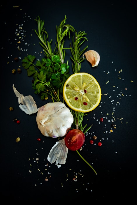 A composition of fresh ingredients, including a whole garlic bulb, a lemon slice, rosemary sprigs, parsley leaves, a small tomato, and scattered peppercorns and salt on a dark background.
