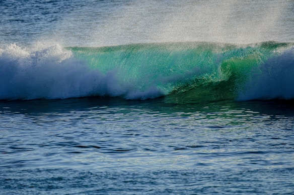 A vibrant turquoise wave crashes in the ocean, with sunlight illuminating the water and creating a misty spray above the crest. The surface of the surrounding sea appears calm yet textured, with various shades of blue.