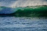 A dynamic ocean wave with a vibrant green hue curling over itself, topped with a frothy white crest. Sunlight is illuminating the wave, creating a translucent effect, while the water appears textured and detailed.