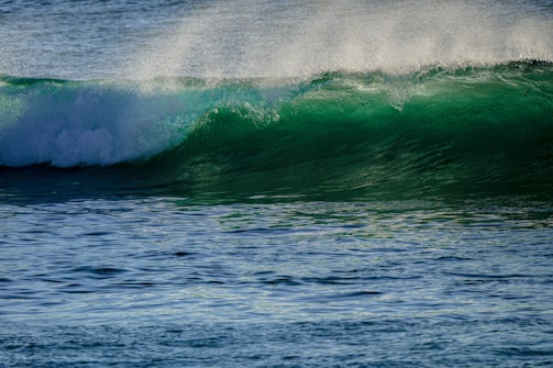 A dynamic ocean wave with a vibrant green hue curling over itself, topped with a frothy white crest. Sunlight is illuminating the wave, creating a translucent effect, while the water appears textured and detailed.