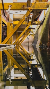An industrial structure features yellow metal beams and pipes forming geometric patterns, with reflections on a still surface that mirror the intricate design above. The beams and framework create a sense of depth and complexity.