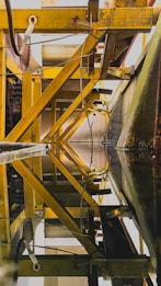 An industrial structure features yellow metal beams and pipes forming geometric patterns, with reflections on a still surface that mirror the intricate design above. The beams and framework create a sense of depth and complexity.