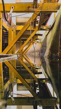 An industrial structure features yellow metal beams and pipes forming geometric patterns, with reflections on a still surface that mirror the intricate design above. The beams and framework create a sense of depth and complexity.