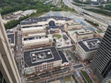 Aerial view of a sprawling urban development with a mix of commercial and residential buildings. Central open space with a circular design, surrounded by brick facades and multiple streets with cars and greenery. Highway visible in the distance along with a dense tree line.