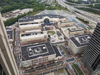 Aerial view of a sprawling urban development with a mix of commercial and residential buildings. Central open space with a circular design, surrounded by brick facades and multiple streets with cars and greenery. Highway visible in the distance along with a dense tree line.