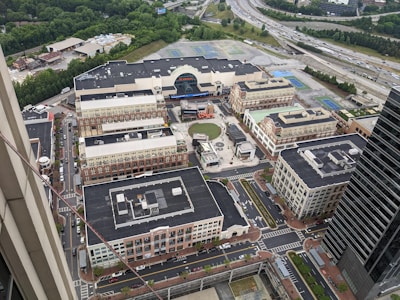 Aerial view of a sprawling urban development with a mix of commercial and residential buildings. Central open space with a circular design, surrounded by brick facades and multiple streets with cars and greenery. Highway visible in the distance along with a dense tree line.