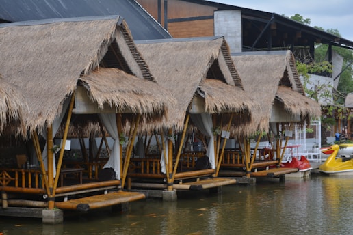A row of bamboo huts with thatched roofs sits by the edge of a tranquil body of water. Each hut is equipped with open seating areas with tables inside, and white curtains hanging from the sides. The huts are supported on stilts over the water, and there are colorful paddle boats in the shape of ducks nearby. The background shows an industrial-style building with a mix of wood and concrete elements.