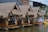 A row of bamboo huts with thatched roofs sits by the edge of a tranquil body of water. Each hut is equipped with open seating areas with tables inside, and white curtains hanging from the sides. The huts are supported on stilts over the water, and there are colorful paddle boats in the shape of ducks nearby. The background shows an industrial-style building with a mix of wood and concrete elements.