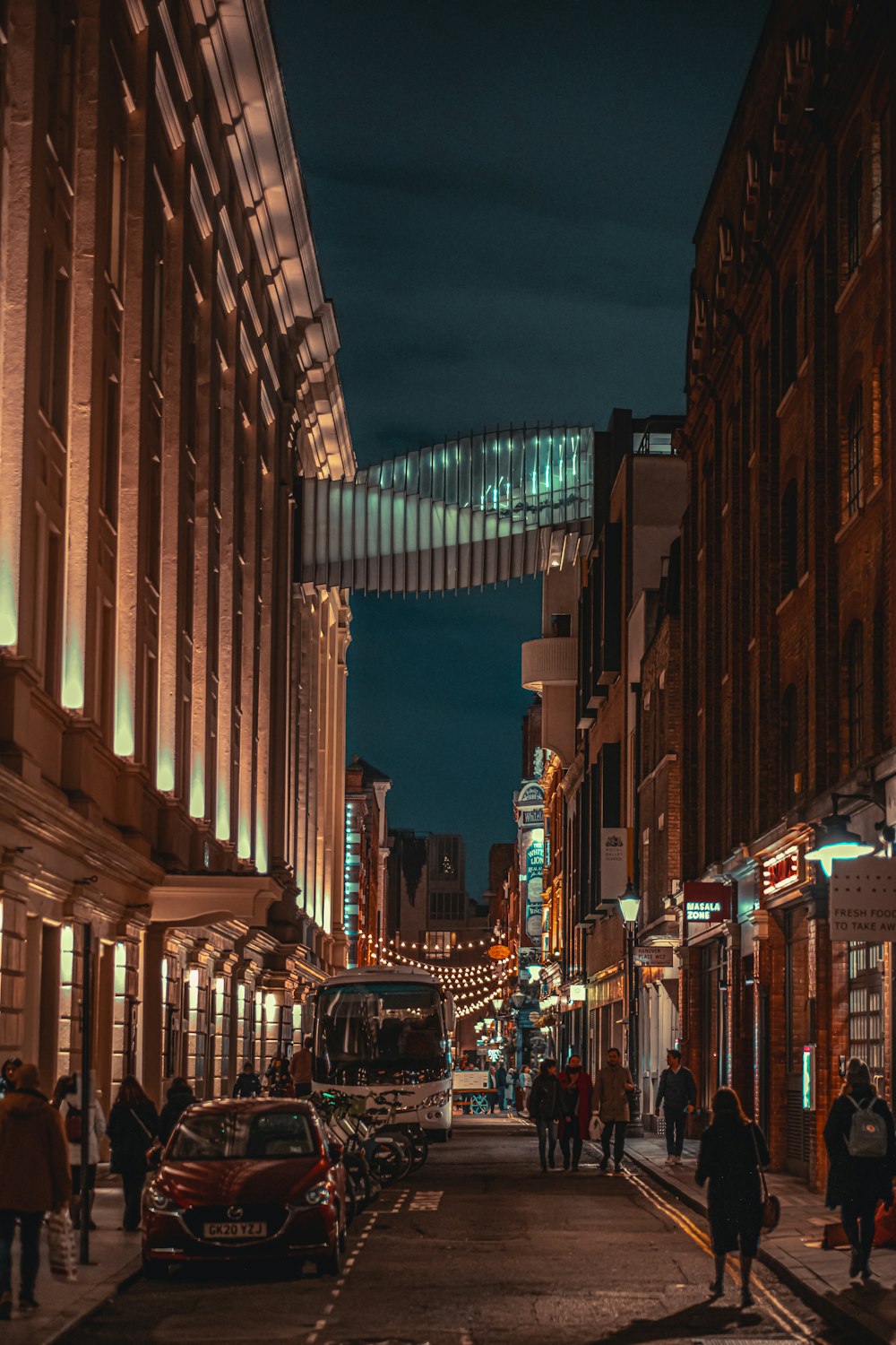 una calle de la ciudad por la noche con gente caminando y coches aparcados a un lado de la