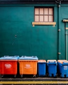 Four large waste bins in bright orange and blue colors are aligned against a dark green wall. Above them is a small rectangular window with wooden framing. The bins are placed on a paved surface marked with yellow lines.