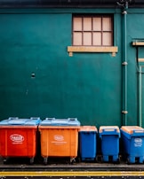 Four large waste bins in bright orange and blue colors are aligned against a dark green wall. Above them is a small rectangular window with wooden framing. The bins are placed on a paved surface marked with yellow lines.