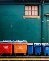 Four large waste bins in bright orange and blue colors are aligned against a dark green wall. Above them is a small rectangular window with wooden framing. The bins are placed on a paved surface marked with yellow lines.