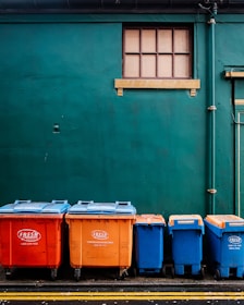 Four large waste bins in bright orange and blue colors are aligned against a dark green wall. Above them is a small rectangular window with wooden framing. The bins are placed on a paved surface marked with yellow lines.