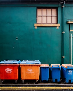 Four large waste bins in bright orange and blue colors are aligned against a dark green wall. Above them is a small rectangular window with wooden framing. The bins are placed on a paved surface marked with yellow lines.