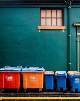 Four large waste bins in bright orange and blue colors are aligned against a dark green wall. Above them is a small rectangular window with wooden framing. The bins are placed on a paved surface marked with yellow lines.