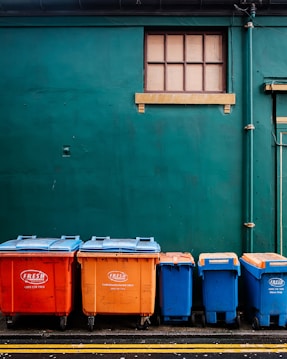 Four large waste bins in bright orange and blue colors are aligned against a dark green wall. Above them is a small rectangular window with wooden framing. The bins are placed on a paved surface marked with yellow lines.