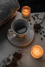 A cup of coffee sits on a saucer surrounded by scattered coffee beans and two lit candles, creating a cozy atmosphere. A folded towel is visible in the background, suggesting a relaxing or spa-like setting.
