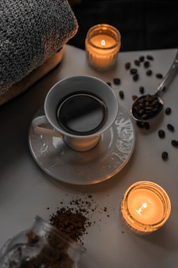 A cup of coffee sits on a saucer surrounded by scattered coffee beans and two lit candles, creating a cozy atmosphere. A folded towel is visible in the background, suggesting a relaxing or spa-like setting.