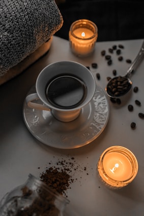 A cup of coffee sits on a saucer surrounded by scattered coffee beans and two lit candles, creating a cozy atmosphere. A folded towel is visible in the background, suggesting a relaxing or spa-like setting.