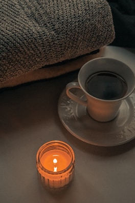 A cozy setup featuring a lit candle in a glass jar, a cup filled with a dark beverage on a saucer, and a textured knitted blanket. The dim lighting and warm glow from the candle create an intimate and relaxing atmosphere.