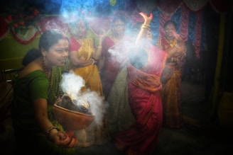 A group of women in traditional attire, engaged in a cultural or religious ritual indoors. One woman is holding a vessel with smoke rising from it, while another appears to be dancing or performing a ritualistic gesture. They are surrounded by colorful decorations, including drapes and flowers in shades of red, yellow, and blue.