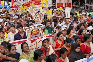 A large group of people are gathered outdoors, many of them holding colorful placards featuring images and writings associated with a cultural or religious event. The participants are dressed in a variety of colorful traditional and modern attire, suggesting a festive or celebratory occasion. Some people are seen wearing hats and colorful sashes, adding to the festive atmosphere.