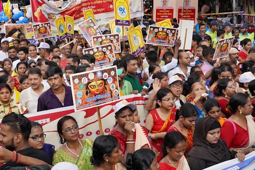 A large group of people are gathered outdoors, many of them holding colorful placards featuring images and writings associated with a cultural or religious event. The participants are dressed in a variety of colorful traditional and modern attire, suggesting a festive or celebratory occasion. Some people are seen wearing hats and colorful sashes, adding to the festive atmosphere.