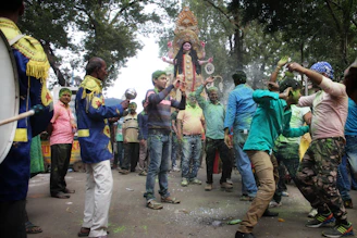 A group of people are gathered on a street surrounded by trees, engaged in a festive celebration. They are dancing energetically, with colored powder visible on their clothes and bodies. In the background, there's a large decorated statue being carried, and the atmosphere is lively and vibrant.