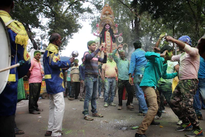 A group of people are gathered on a street surrounded by trees, engaged in a festive celebration. They are dancing energetically, with colored powder visible on their clothes and bodies. In the background, there's a large decorated statue being carried, and the atmosphere is lively and vibrant.
