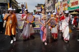 A group of people is participating in a street event. One person in the center is wearing vibrant and traditional clothing, adorned with a deity-inspired costume, holding a prop depicting a snake. Others are dressed in traditional sarees and white caps, carrying colorful circular fans. Banners with text are visible in the background, suggesting an organized parade or cultural celebration.
