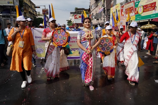 A group of people is participating in a street event. One person in the center is wearing vibrant and traditional clothing, adorned with a deity-inspired costume, holding a prop depicting a snake. Others are dressed in traditional sarees and white caps, carrying colorful circular fans. Banners with text are visible in the background, suggesting an organized parade or cultural celebration.