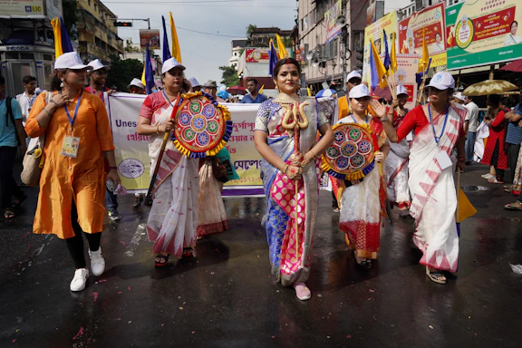 A group of people is participating in a street event. One person in the center is wearing vibrant and traditional clothing, adorned with a deity-inspired costume, holding a prop depicting a snake. Others are dressed in traditional sarees and white caps, carrying colorful circular fans. Banners with text are visible in the background, suggesting an organized parade or cultural celebration.