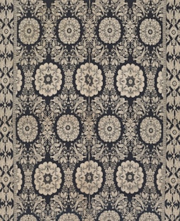 Intricate floral and geometric patterns in symmetrical arrangements dominate the textile design. Cream-colored motifs stand out against a dark background, forming a network of stylized flowers and ornate shapes. The design is bordered by a repetitive pattern of similar motifs.
