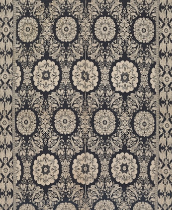 Intricate floral and geometric patterns in symmetrical arrangements dominate the textile design. Cream-colored motifs stand out against a dark background, forming a network of stylized flowers and ornate shapes. The design is bordered by a repetitive pattern of similar motifs.