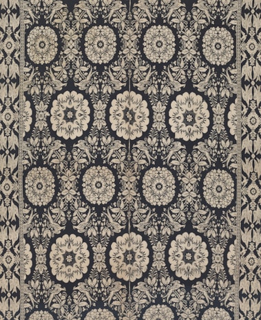 Intricate floral and geometric patterns in symmetrical arrangements dominate the textile design. Cream-colored motifs stand out against a dark background, forming a network of stylized flowers and ornate shapes. The design is bordered by a repetitive pattern of similar motifs.